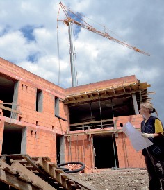 Foto eines Neubaus, mit einem Kran im Hintergrund und einer Frau, welche Baupläne in der Hand hält, im Vordergrund.
